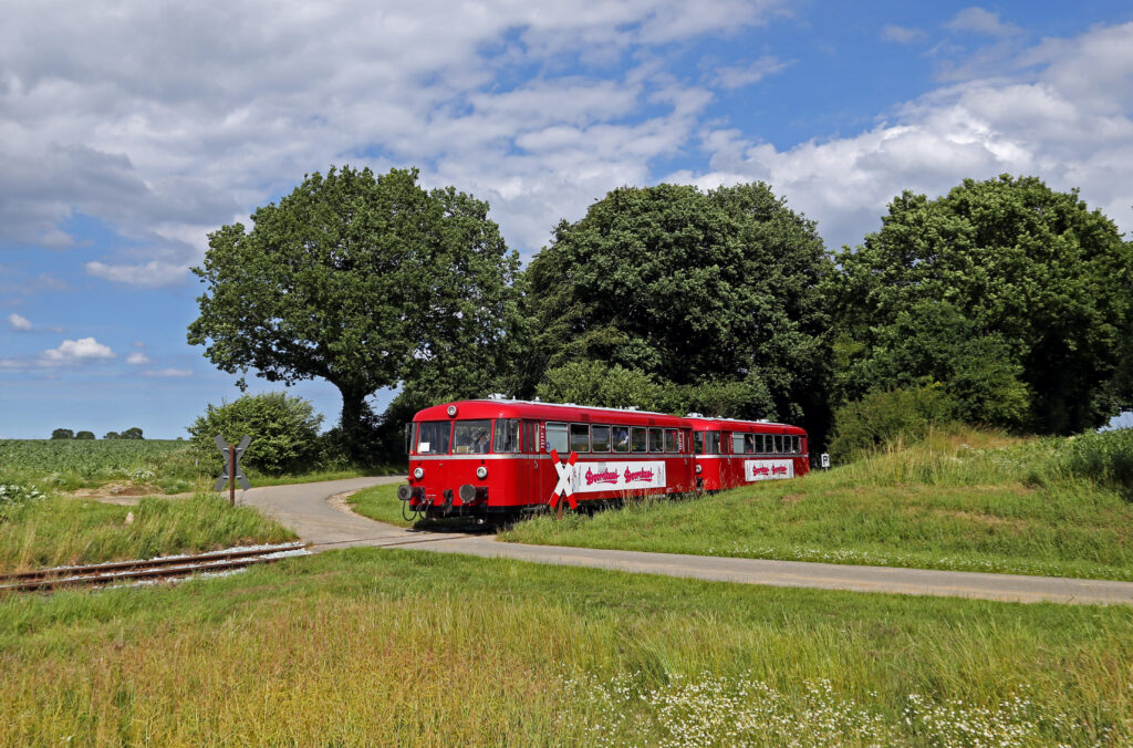 Zwei rote, alte Triebwagen queren einen Bahnübergang. Im Vordergrund eine grüne Wiese, im Hintergrund große Bäume.